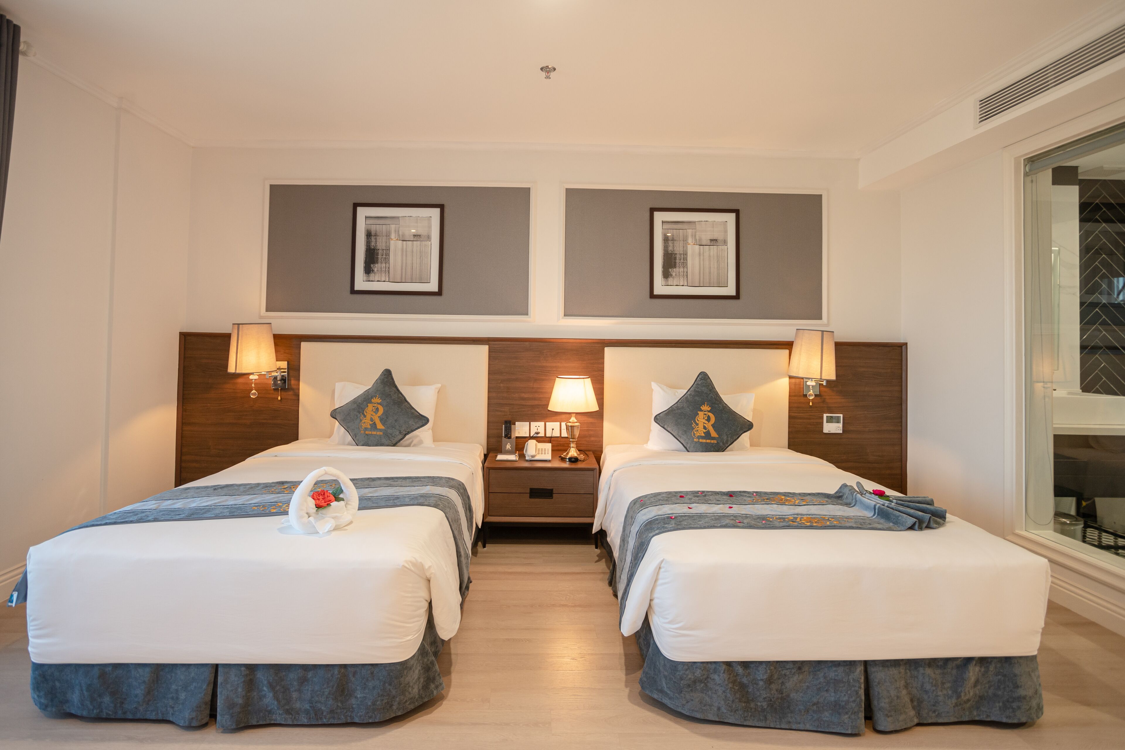 Une chambre d’hôtel avec deux lits, une table de chevet, des lampes fixées au mur et des tableaux encadrés au-dessus des lits.