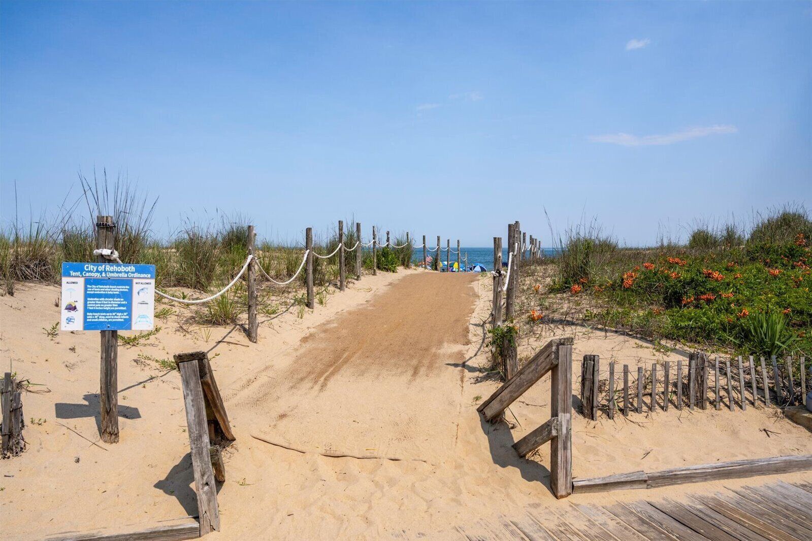 Un sendero de arena que conduce a una pasarela de madera con un letrero que indica las normas de la playa.
