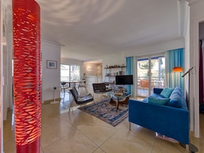 Een moderne woonkamer met een blauwe bank, een eethoek met stoelen, een keuken met een televisie en een groot raam met gordijnen.