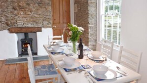 A dining area with a stone wall, a fireplace, and a table set for four with white chairs. - Rose Cottage (St Austell)