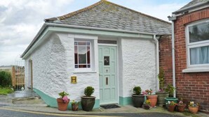 A small, single-story building with a white exterior and a green door. There are several potted plants in front of the building. - Rose Cottage (St Austell)