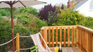 A wooden deck with a rope barrier, a striped hammock, a green chair, a small round table, and a bottle of champagne. - Rose Cottage (St Austell)