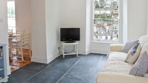 A living room with a dark tiled floor, a white sofa with striped cushions, a television on a white stand, and a window with a city view. - Rose Cottage (St Austell)