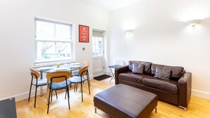A modern living room with a brown leather sofa, a dining table with chairs, and a window with a view of trees. - Bright & Comfortable Angel Apartment (London)