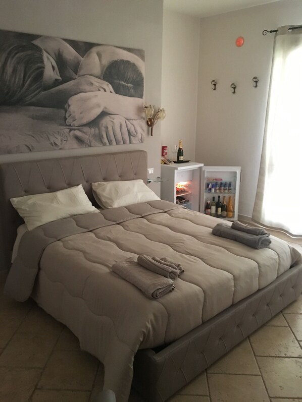 Ein Schlafzimmer mit einem großen Bett, einem Kopfteil mit einem Gemälde, einer Minibar und einem Fenster mit Vorhängen.