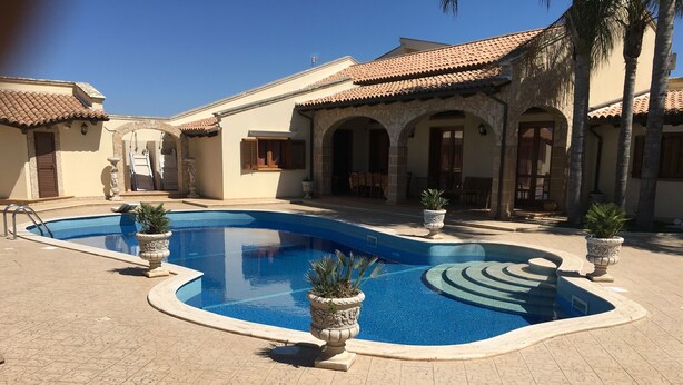 A swimming pool with a curved design, surrounded by a tiled patio and potted plants. - "FORTUNA" Luxury double room in prestigious Villa with shared pool. (Mazara del Vallo)