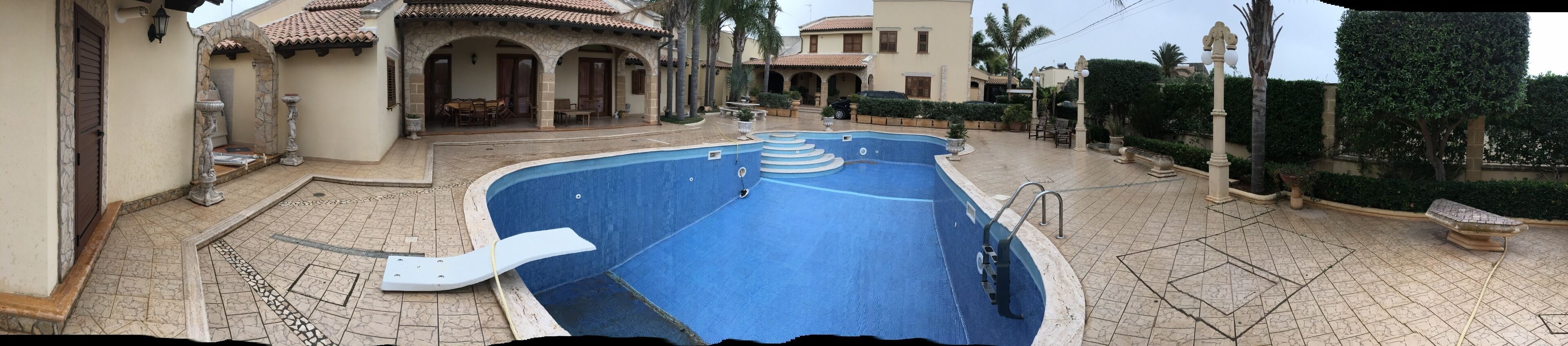 Ein Pool mit Stein оформлением, eine überdachte Terrasse und ein Gebäude mit einem Ziegeldach und Bögen.
