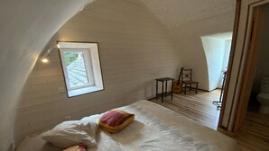 Un dormitorio con techo inclinado, una cama con ropa de cama blanca, una guitarra, una silla y una mesita.