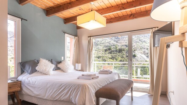 A bedroom with a bed, bench, bedside table, and a view of the outdoors. - VC2 - LUXURY 2 BEDROOM APARTMENT CERRO CATEDRAL (San Carlos de Bariloche)