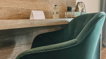 Une chambre d’hôtel moderne avec un plancher en bois, un fauteuil recouvert d’un tissu vert, un téléviseur à écran plat fixé au mur, un bureau en bois avec un menu, une bouteille d’eau et un verre.