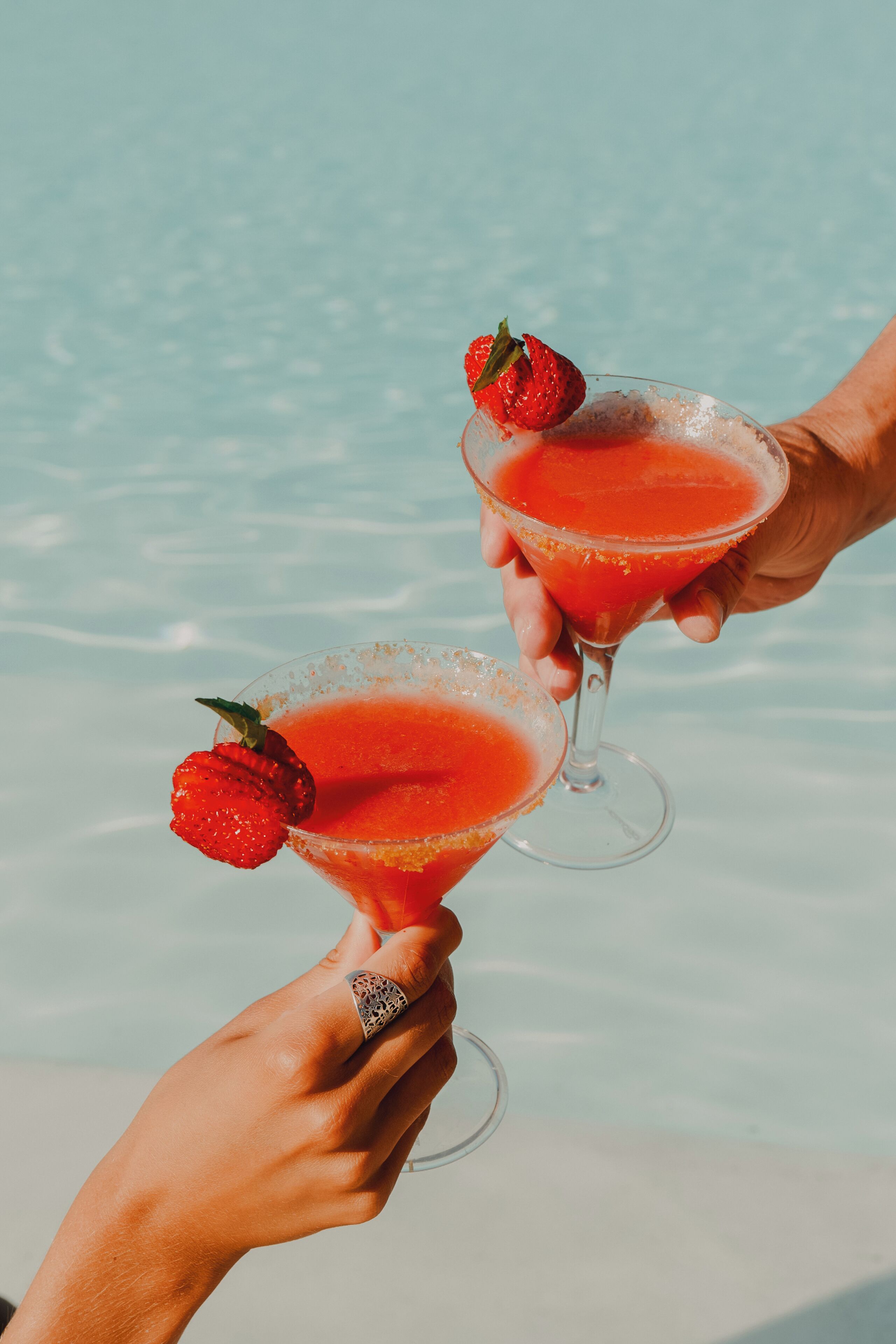 Deux mains tenant des verres à martini remplis d’une boisson rouge et décorés de fraises.
