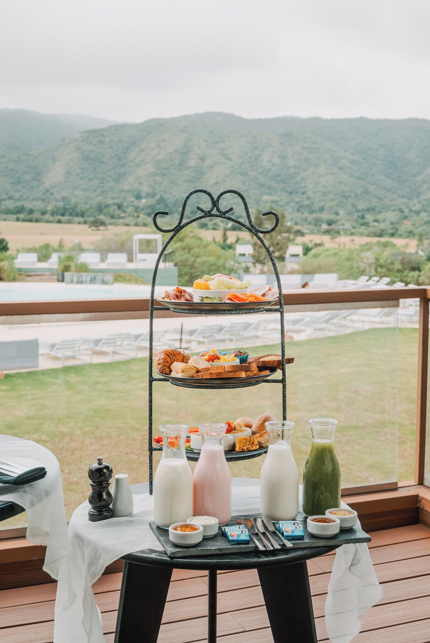 Un buffet per la colazione con diverse pietanze e bevande su un espositore a più livelli, sistemato su un pontile di legno con vista panoramica sulle montagne.