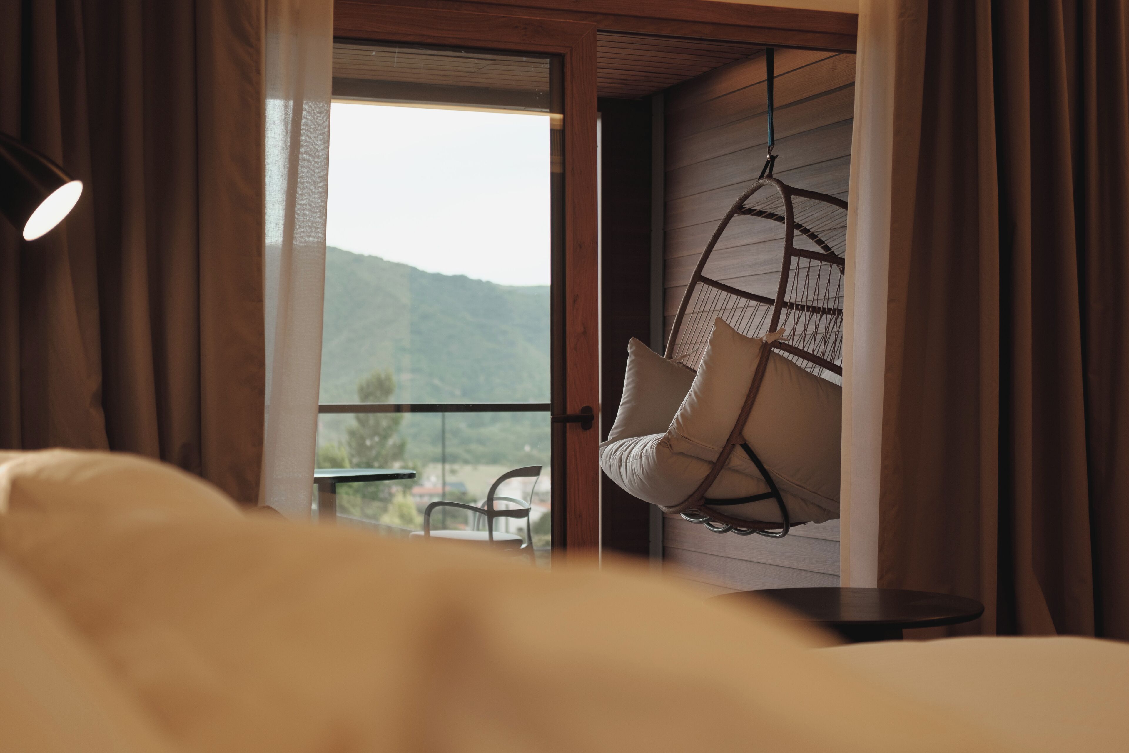 Une chambre avec un lit, un fauteuil suspendu et un balcon offrant une vue sur les collines.
