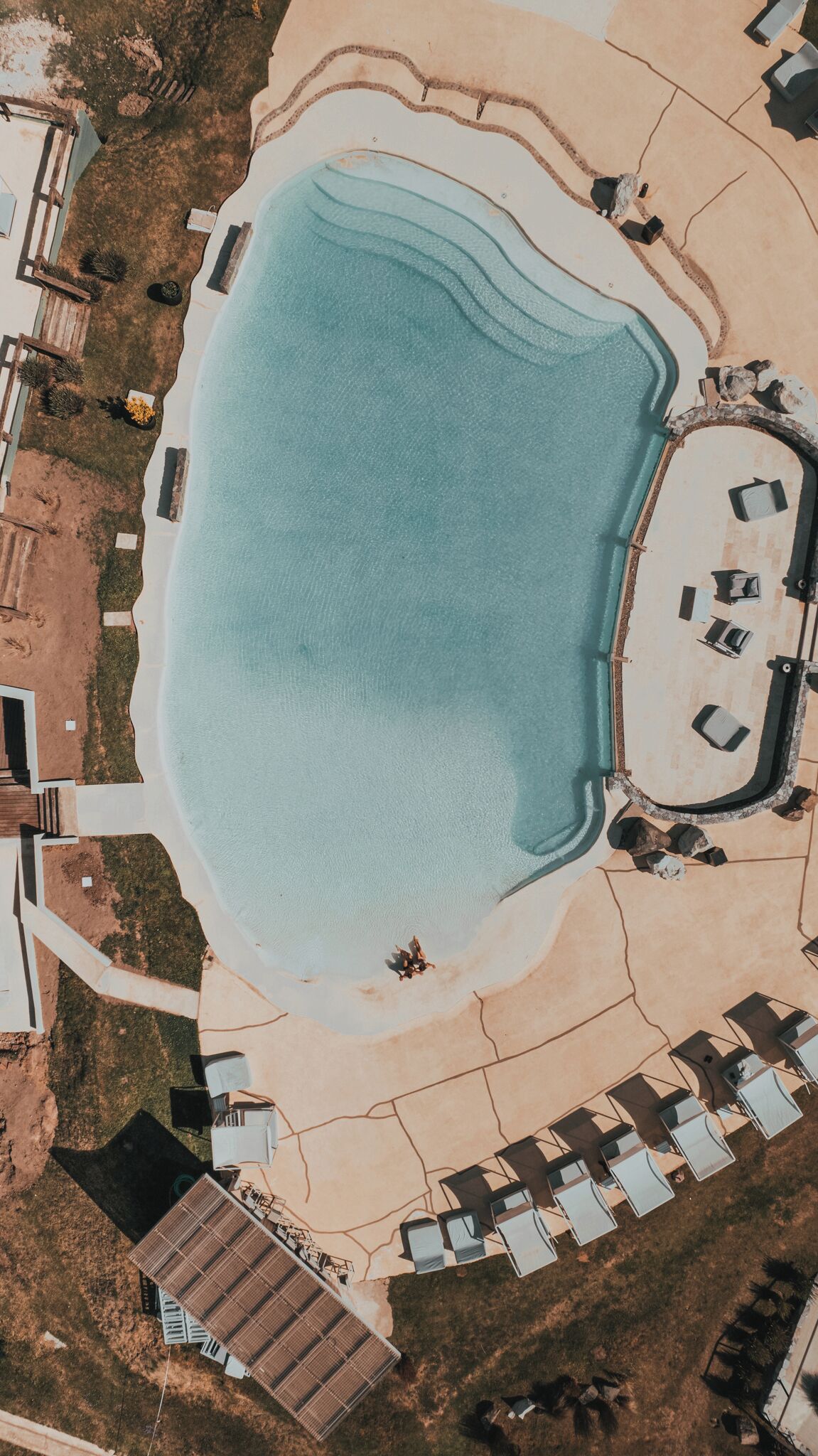 Vue aérienne d’un complexe hôtelier doté d’une grande piscine, de transats et d’un petit bâtiment.
