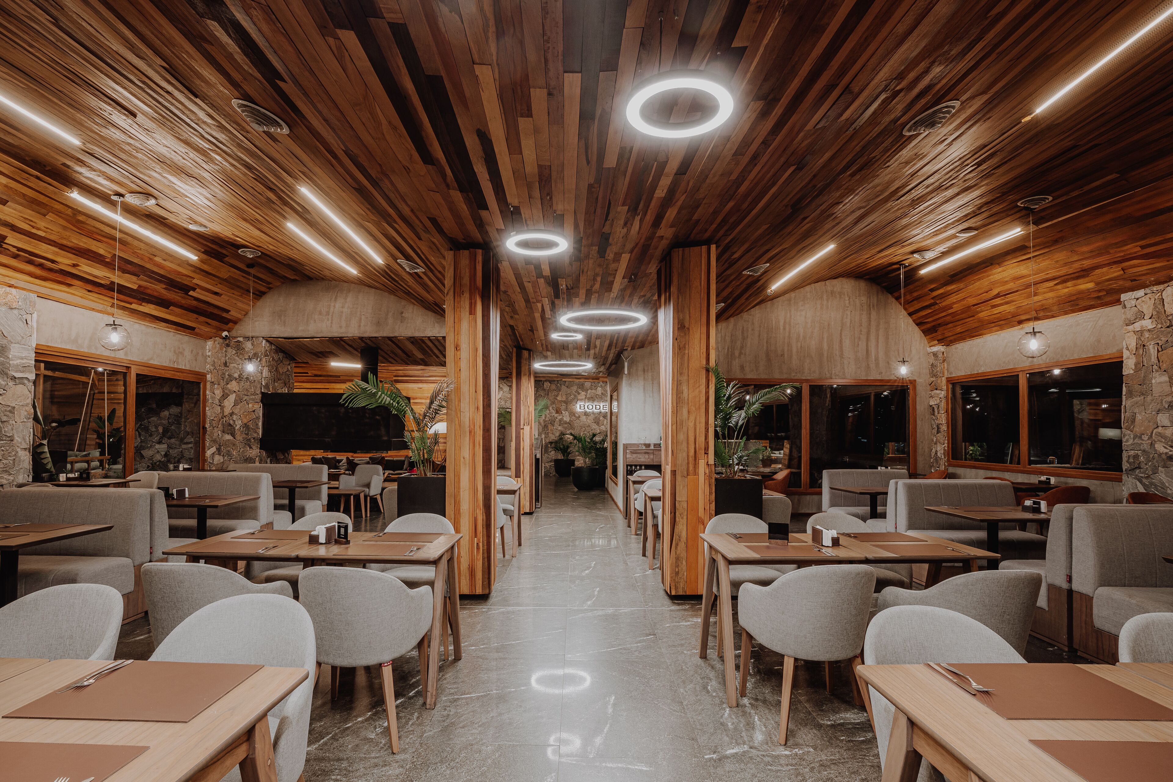 A restaurant with wooden ceiling panels, circular ceiling lights, and stone walls. Tables and chairs are arranged for dining.