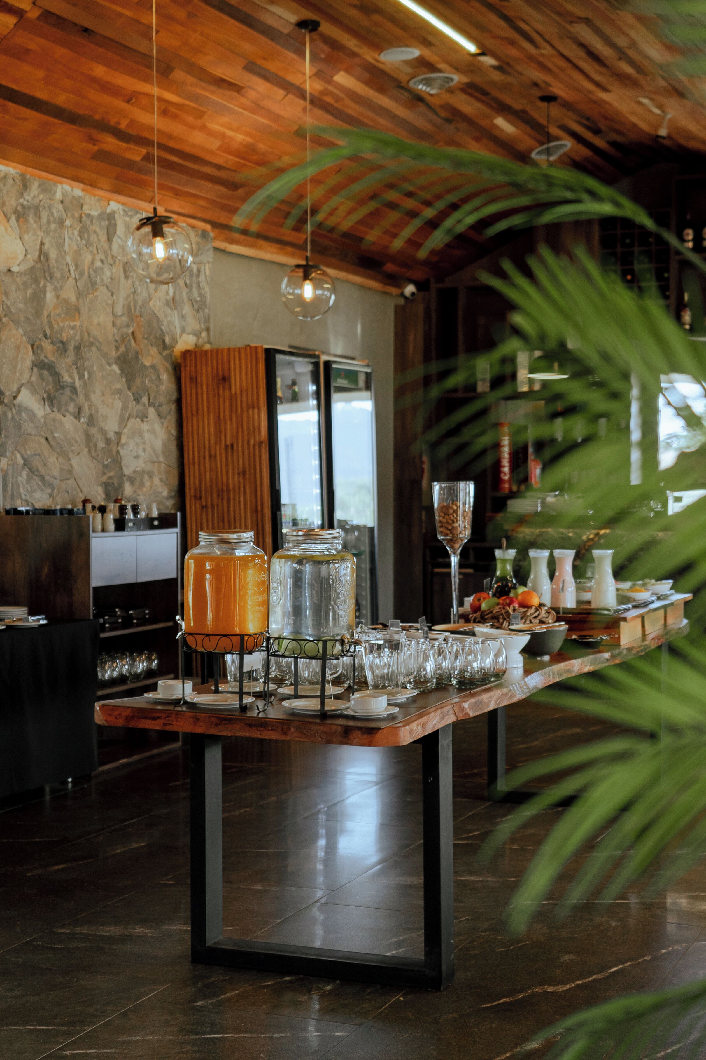 A long dining table with glassware, a fruit bowl, and a beverage dispenser in a modern interior with wooden ceiling and stone wall.