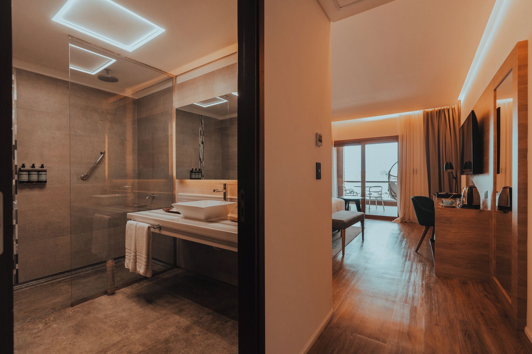 Un baño moderno con ducha envuelta en vidrio, tocador doble y un espejo grande. La habitación tiene piso de madera y vistas al exterior a través de un ventanal amplio.