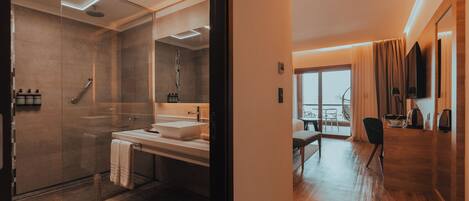 Un baño moderno con ducha envuelta en vidrio, tocador doble y un espejo grande. La habitación tiene piso de madera y vistas al exterior a través de un ventanal amplio.