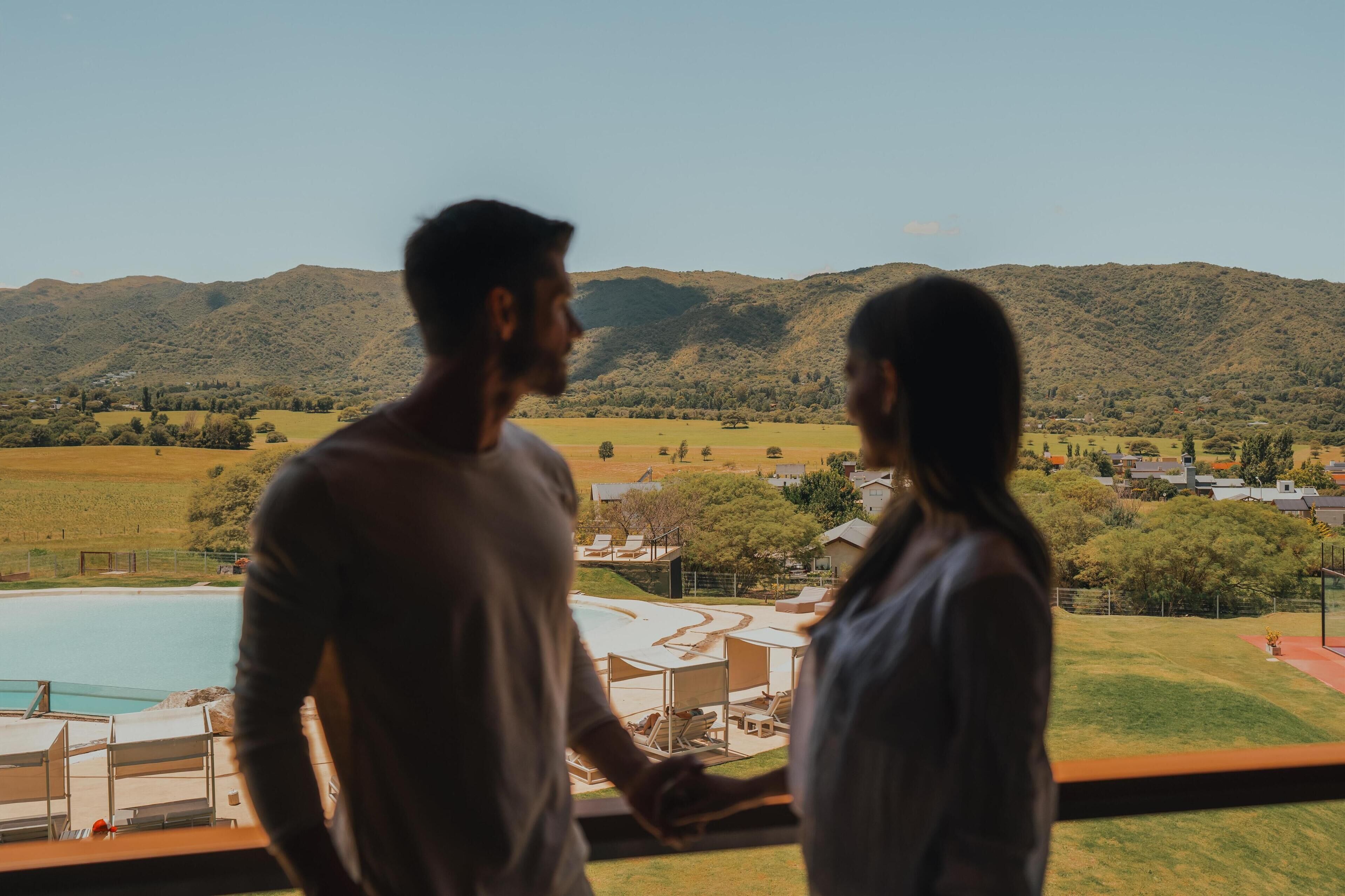 A couple standing by a window overlooking a pool and a mountainous landscape.