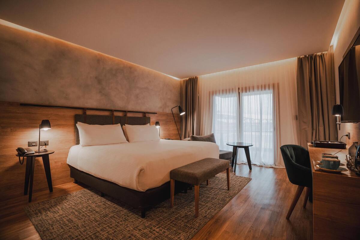 Een moderne hotelkamer met een groot bed, nachtlampjes, een klein tafeltje, een stoel en een bureau met een lamp.