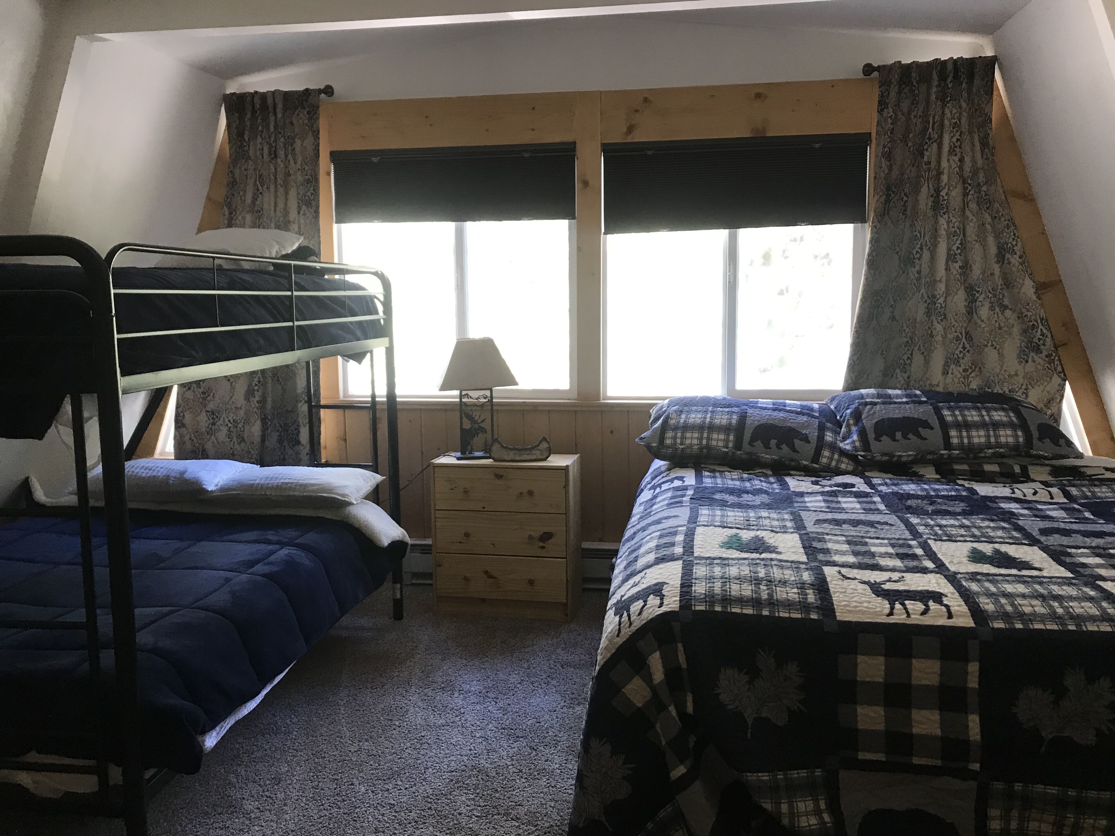 A dormitory room with bunk beds, a bedside table with a lamp, a window with curtains, and a bed with a patterned blanket.