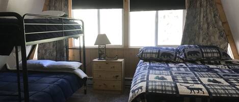 A dormitory room with bunk beds, a bedside table with a lamp, a window with curtains, and a bed with a patterned blanket.