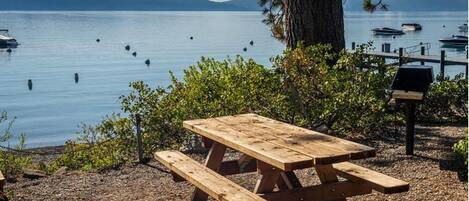 Una mesa de picnic de madera con bancos, rodeada de árboles y una vista de un lago tranquilo con barcos.