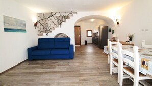 Ein Wohnzimmer mit blauem Sofa, Holzboden, einem Esstisch mit Stühlen und einer Küche im Hintergrund.