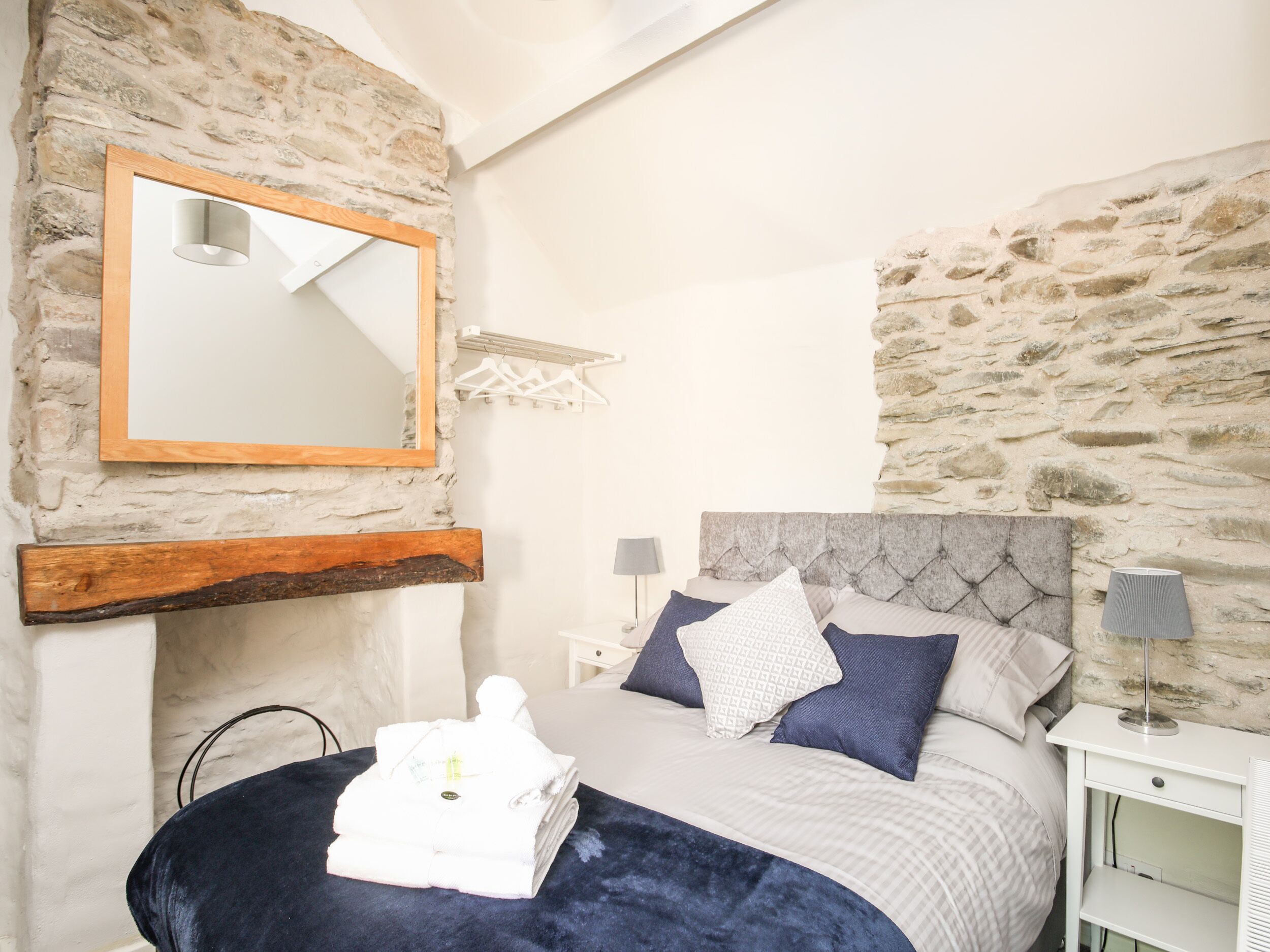 Un dormitorio con pared de piedra, cabecera de madera, cama con manta azul y mesita de noche blanca con lámpara.