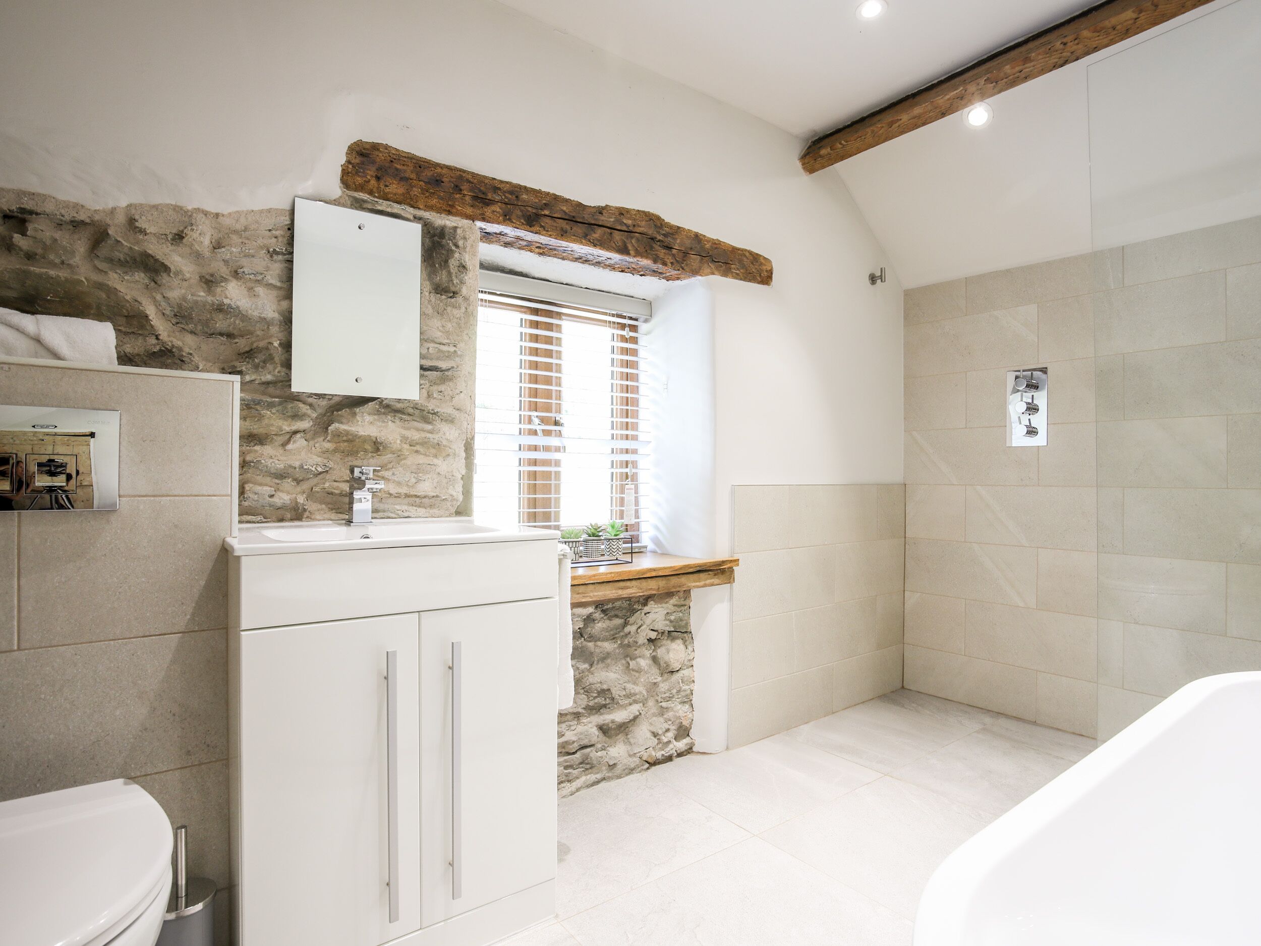A bathroom with a stone wall, a wooden beam ceiling, a white sink, and a shower area.