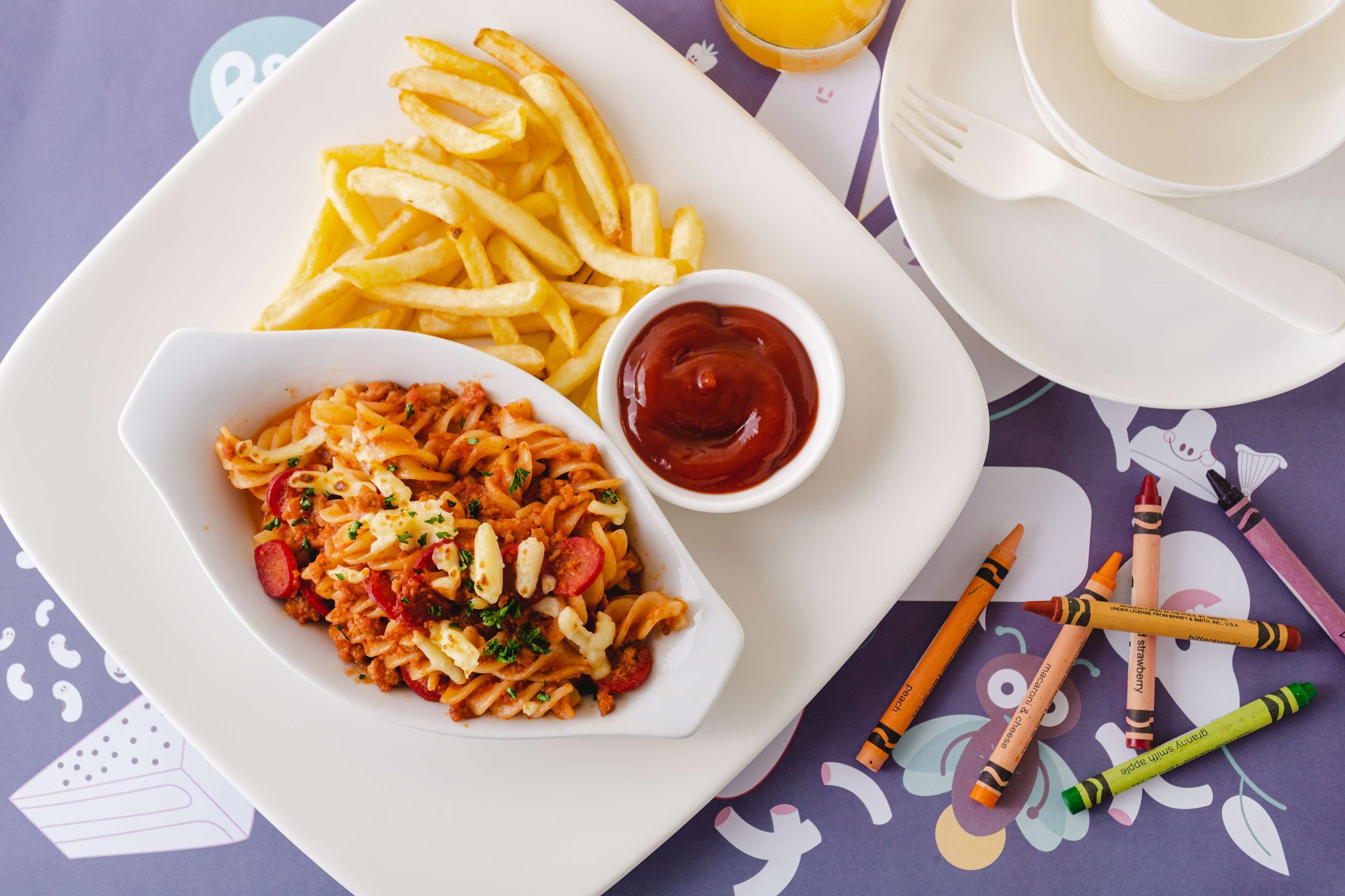 A plate of pasta and fries with ketchup, a glass of orange juice, and a cup of tea on a table with a purple tablecloth and children's crayons.
