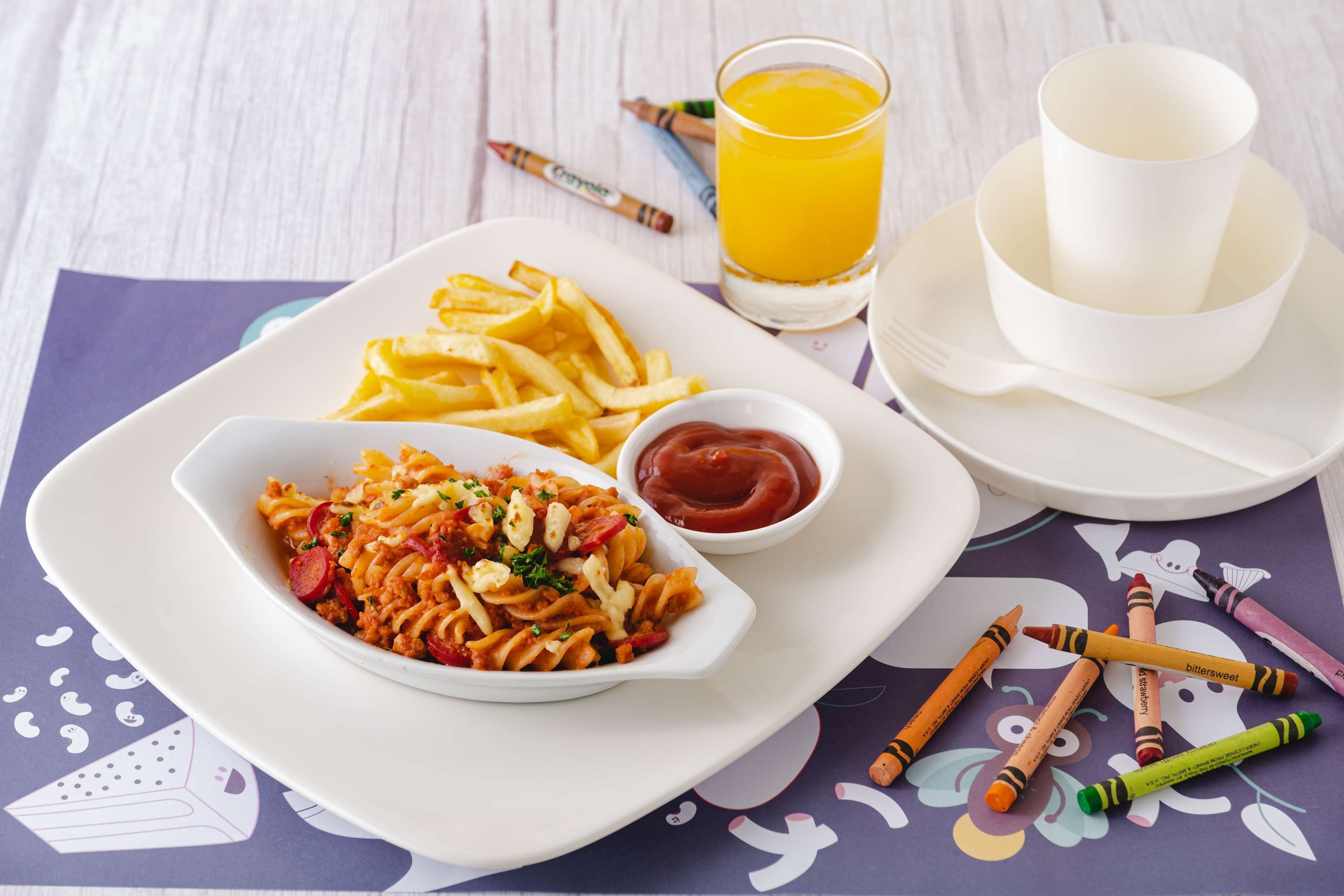 Un plato de pasta con papas fritas, un vaso de jugo de naranja y una taza de café sobre una mesa con crayones.