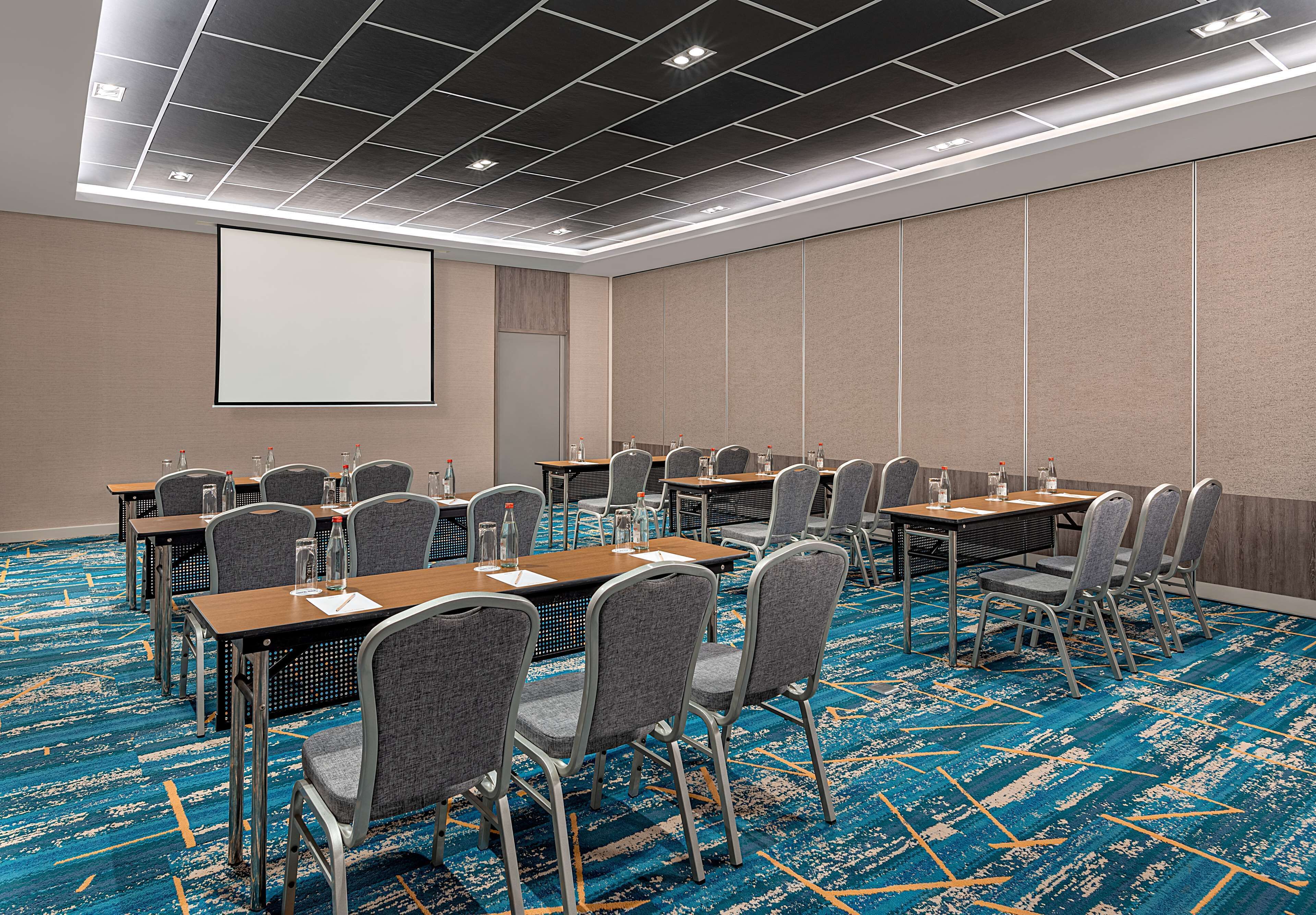 Una sala de conferencias con varias mesas y sillas, una pantalla de proyector y suelo alfombrado.