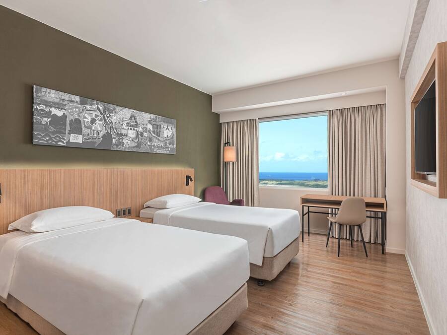 A hotel room with two beds, a desk, a chair, a large window with a view, and a wall-mounted artwork.
