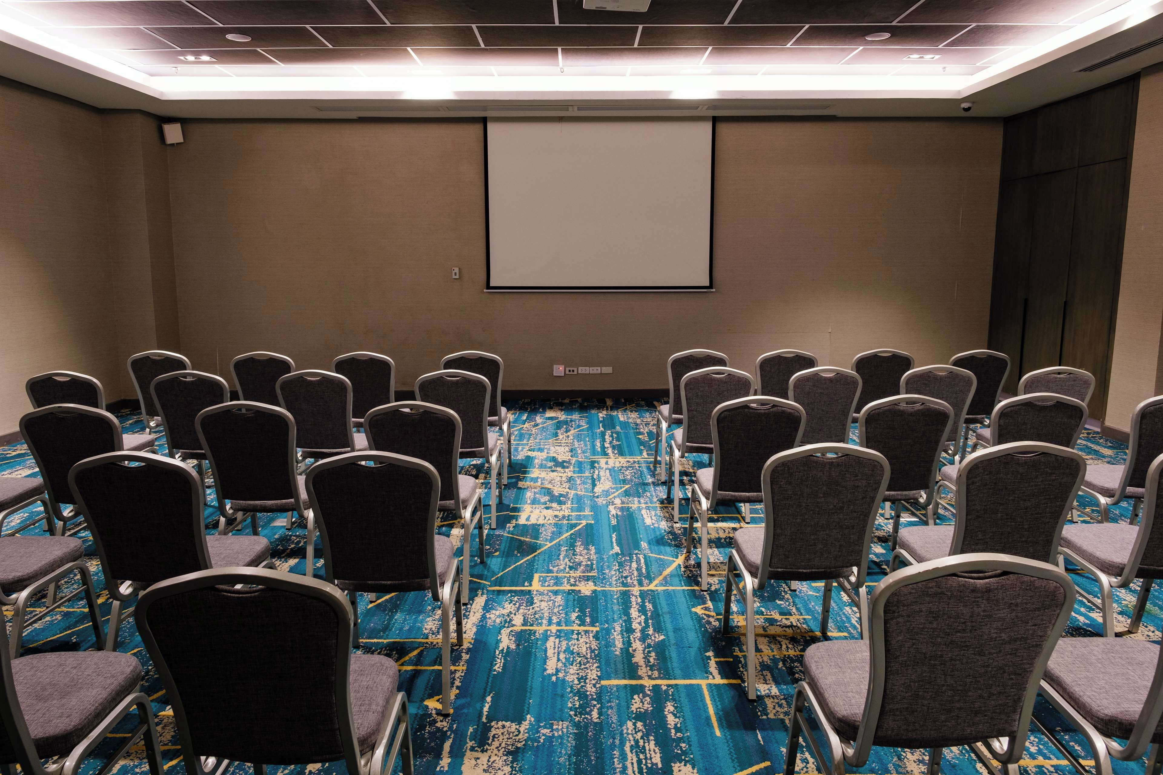 A conference room with rows of chairs facing a projection screen, carpeted floor, and a plain wall.