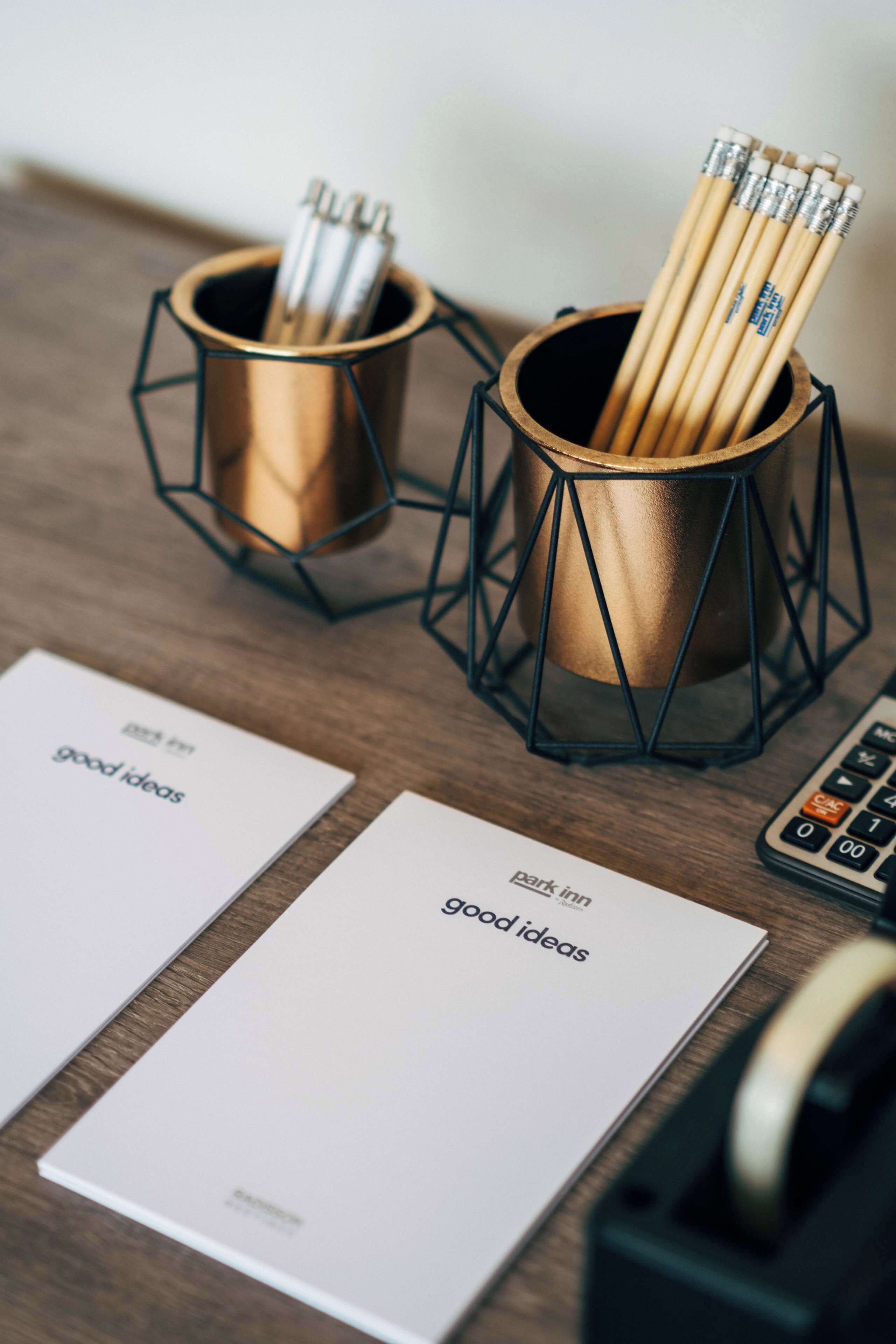 Un escritorio con dos portalápices metálicos geométricos que contienen lápices, una calculadora y dos tarjetas de presentación con el texto 'good ideas'.