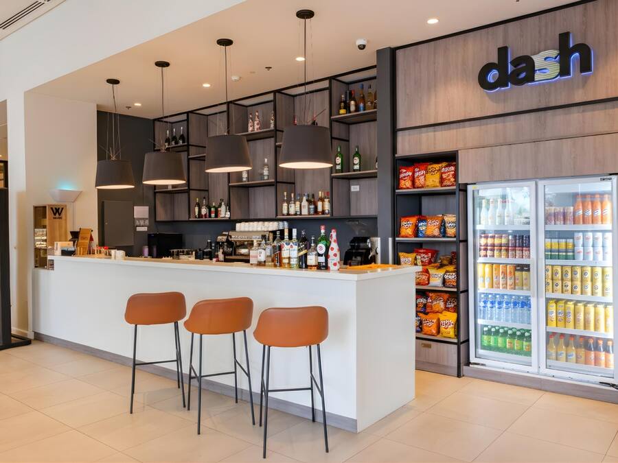 Una zona de bar moderna con mostrador, taburetes, iluminación en el techo, estanterías con botellas y un refrigerador lleno de bebidas.