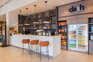 A modern bar area with a counter, stools, overhead lighting, shelves with bottles, and a refrigerator stocked with beverages.