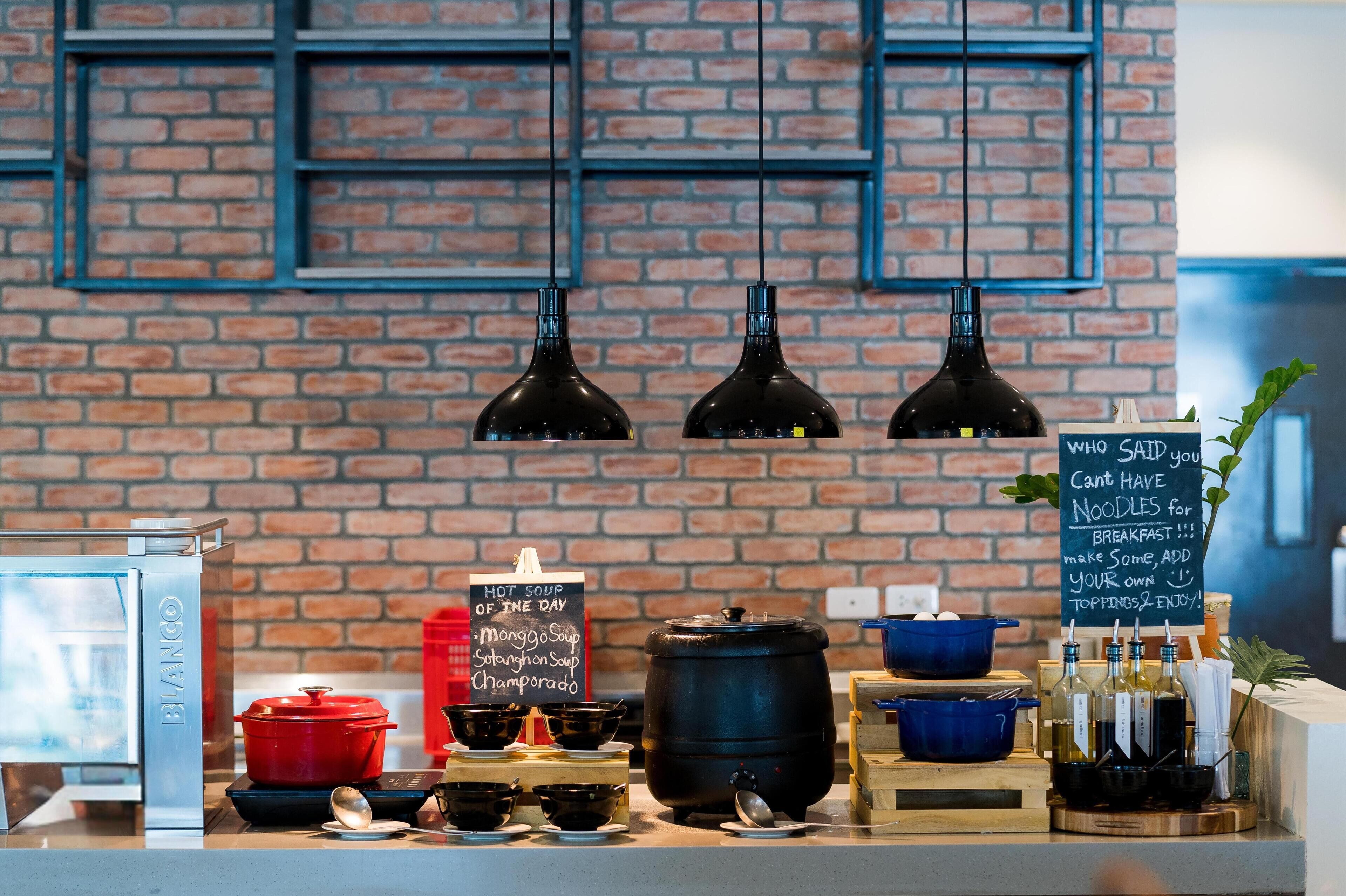 A breakfast bar with a variety of hot soups and beverages, a chalkboard menu, and a brick wall background.