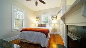 A bedroom with a bed, two bedside lamps, a ceiling fan, a fireplace, and two windows with blinds.