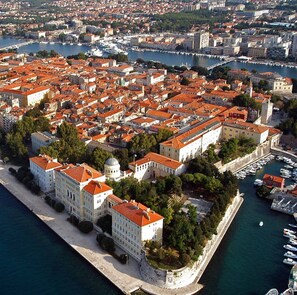 An aerial view of a coastal city with a dense cluster of buildings with red-tiled roofs, a prominent white building with a dome, and a marina with boats. - Obrovackrusevo, Modern apt w. Terrace & sea View (Obrovac)