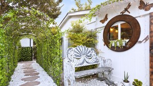 Un chemin de jardin mène à un banc peint en blanc, doté d’un dossier décoratif, et entouré de petits galets blancs et de verdure.