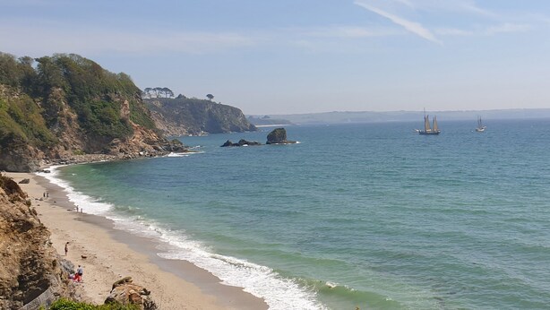 A coastal beach with rocky cliffs, a sandy shore, and sailboats in the distance. - Robin Hill (St Austell)