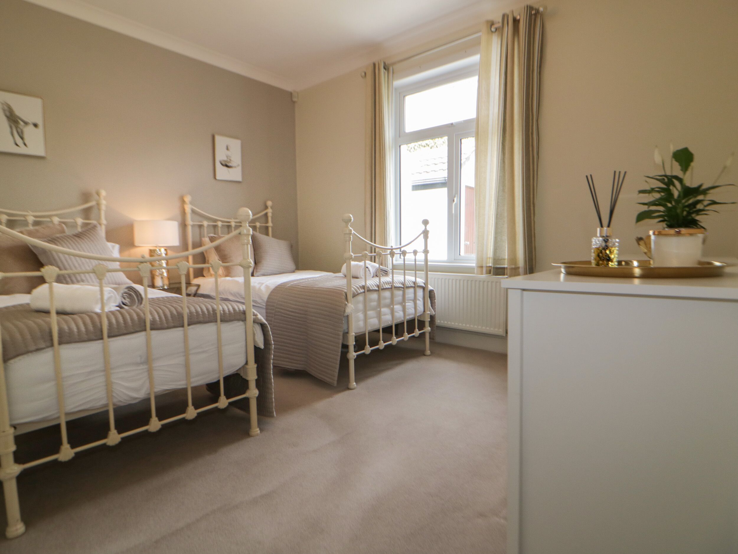 A bedroom with two beds, a window with curtains, a radiator, and a bedside table with a plant and candles.
