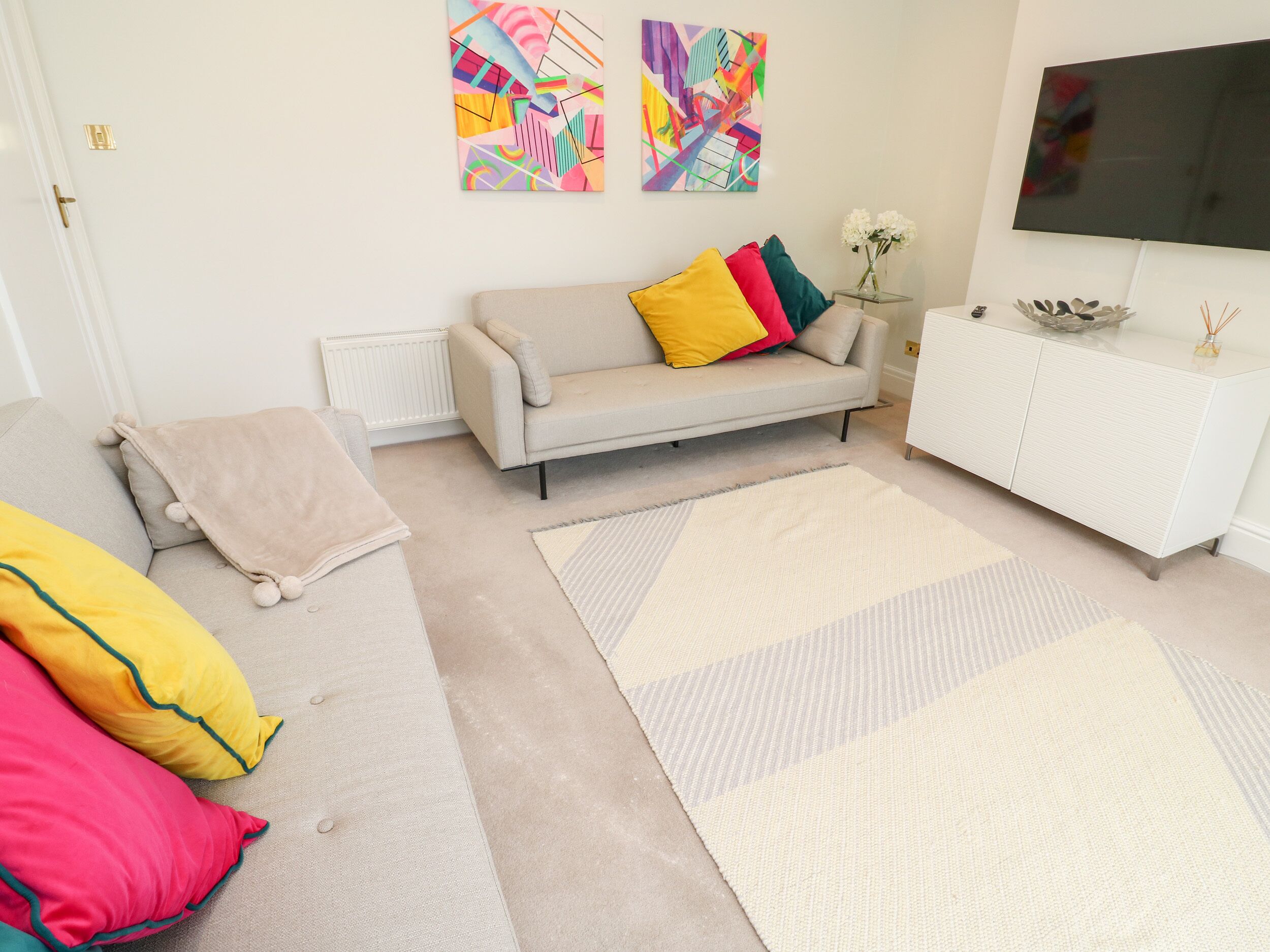 A living room with a sofa, colorful cushions, a large rug, a white cabinet, and abstract paintings on the wall.