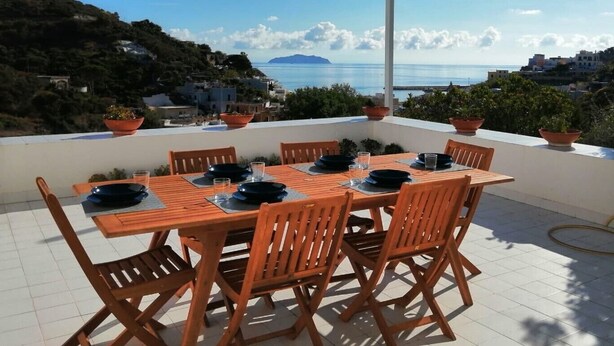 Une table en bois, entourée de chaises, sur une terrasse offrant une vue sur la côte.