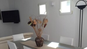 Un salon moderne comprenant une table à manger, des chaises blanches, un canapé beige, un téléviseur à écran plat fixé au mur et un lampadaire.