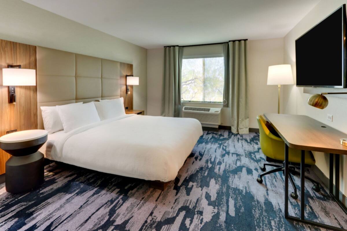Habitación de hotel con una cama grande, un escritorio, una silla, un televisor de pantalla plana, una lámpara y una ventana con cortinas.
