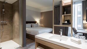 A modern hotel bathroom with a shower, a large mirror, a sink, and a bedside table with a lamp. - AC Hotel by Marriott Wuerzburg (W ürzburg)