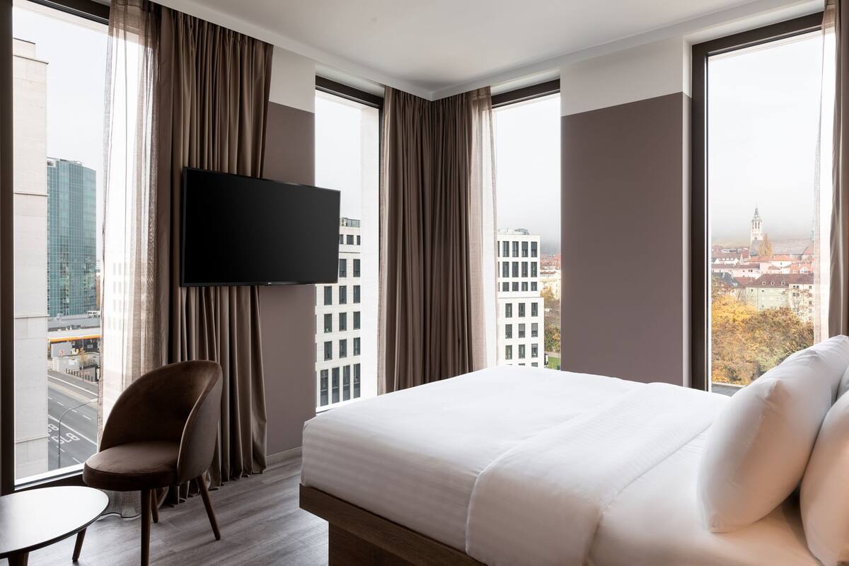 A hotel room with a large bed, a TV mounted on the wall, a chair, and a view of the cityscape through large windows.